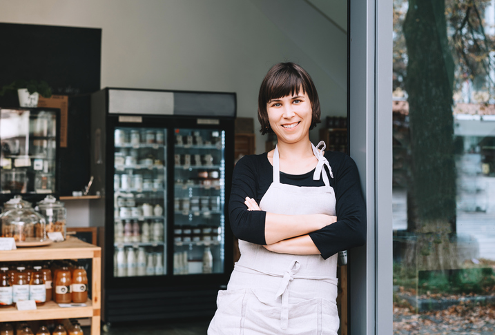 Portrait of owner of sustainable small local business. Shopkeeper of zero waste shop standing on interior background of shop. Smiling young woman in apron welcoming at entrance of plastic free store