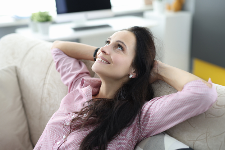 Woman sits on couch with her arms folded behind her head and looks up dreamily. Positive affirmations to lift your mood concept